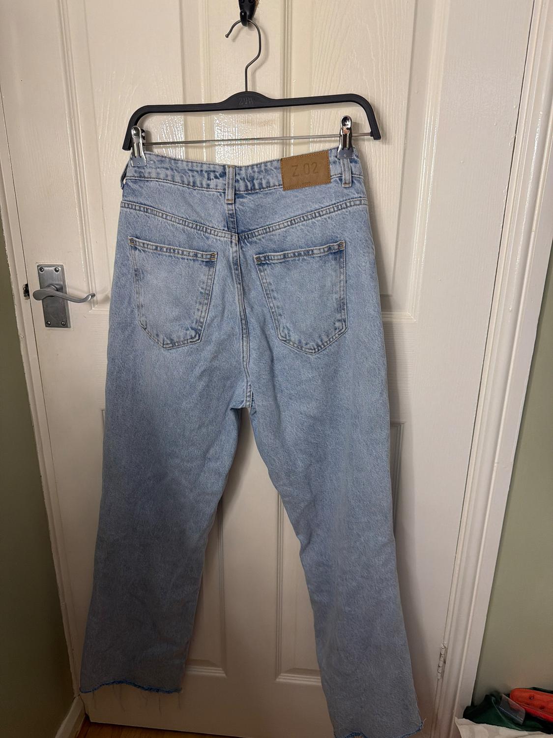 Blue jeans hanging on a black hanger over a white door. Brown tag with "Z.02" text visible on the waistband.