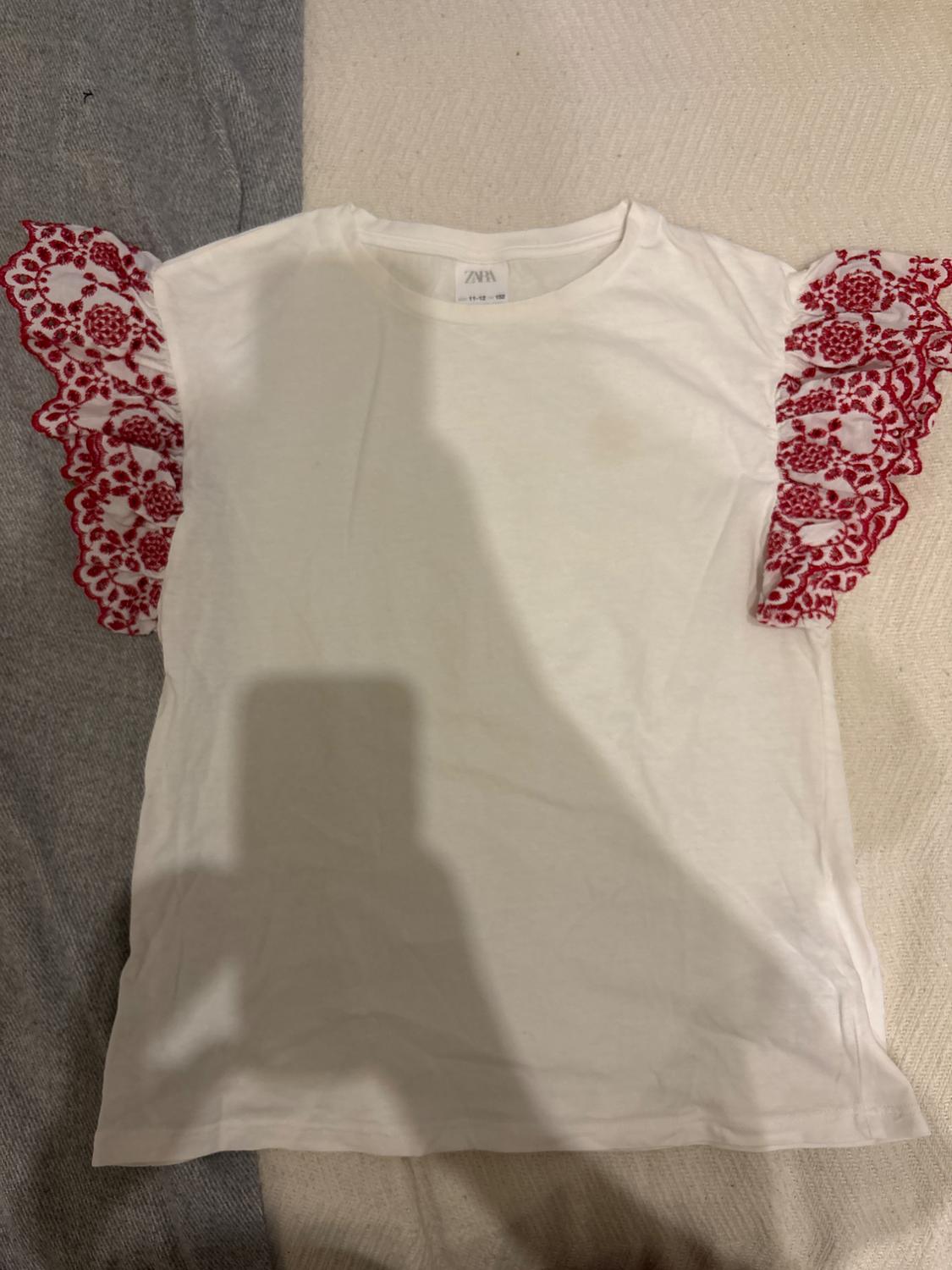 White T-shirt with red lace sleeves on a grey and beige background. Label visible on the neck. Shadow of a phone in the centre.