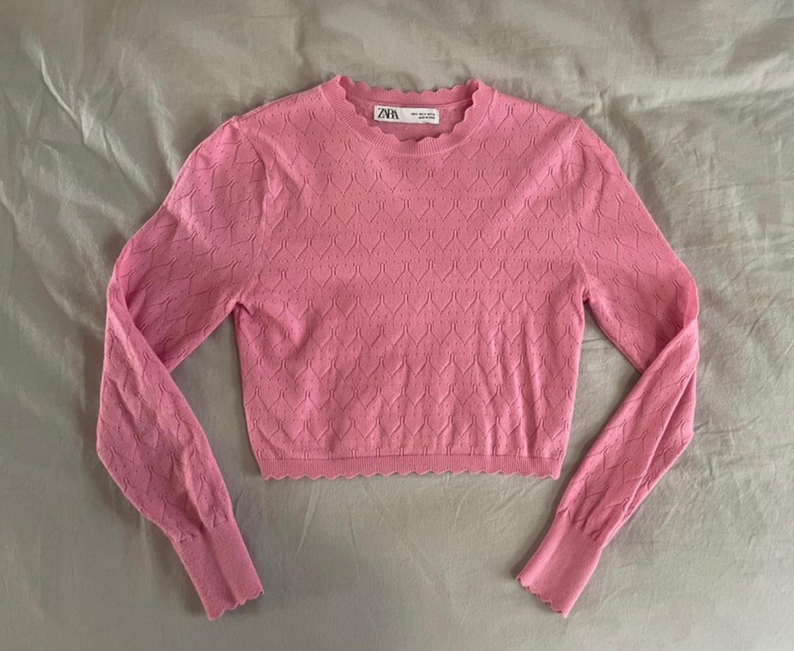 Pink cropped open-knit jumper with long sleeves and scalloped edges over grey fabric. Label bearing the text ZARA.