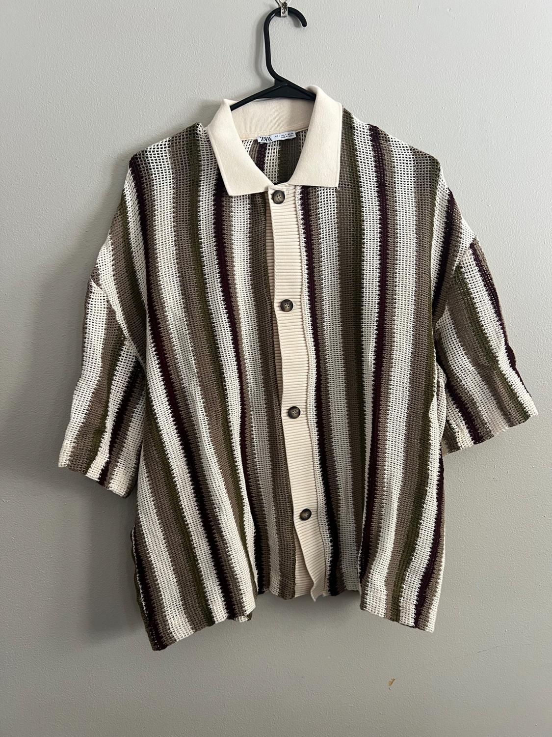 Knit shirt with vertical stripes in beige, brown, and green tones, hanging on a black hanger against a gray wall.