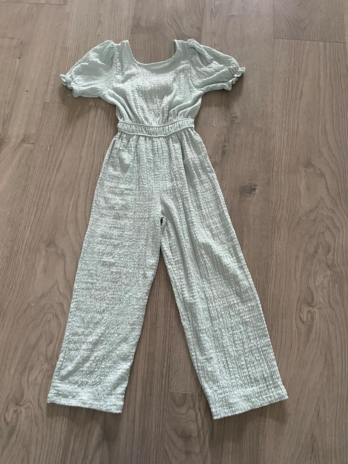 Light-colored textured fabric jumpsuit with puff short sleeves, elastic waist, and wide-leg pants, on a wooden floor.