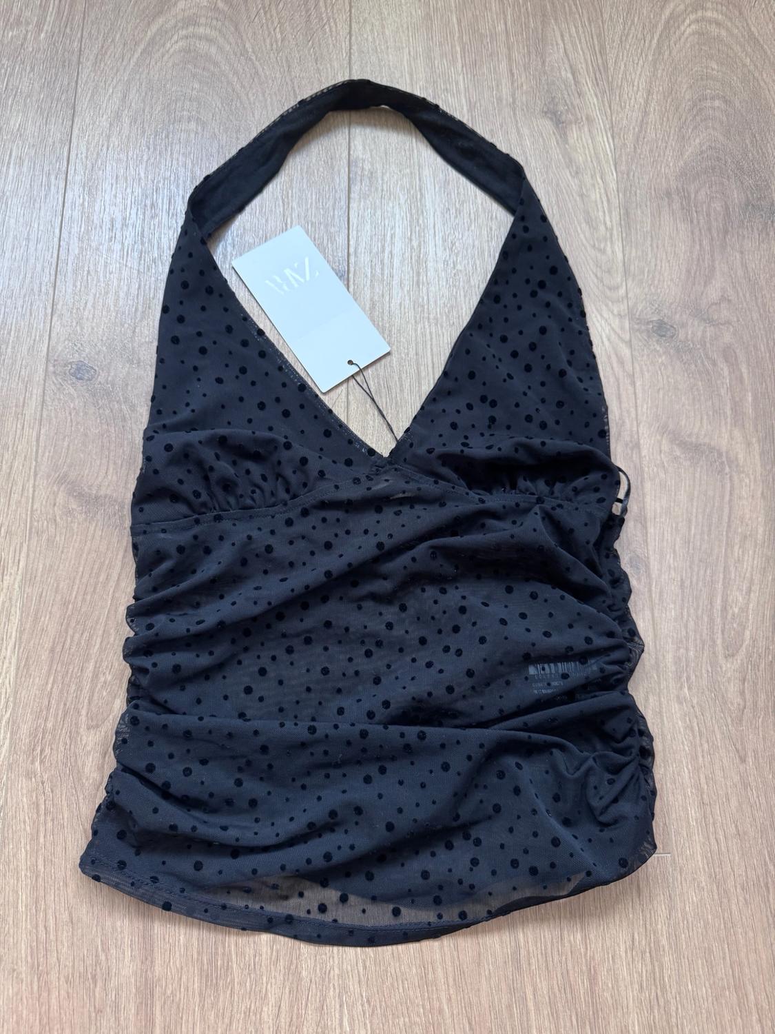 Black halterneck top with a V-neckline, semi-sheer fabric with polka dots and gathers, on a wooden floor. ZARA label.
