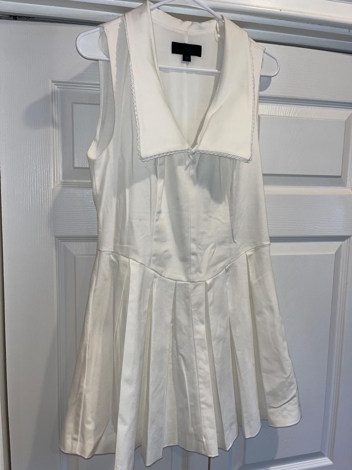Sleeveless white dress with a wide neckline and pleated skirt, hanging on a hanger in front of a white door.