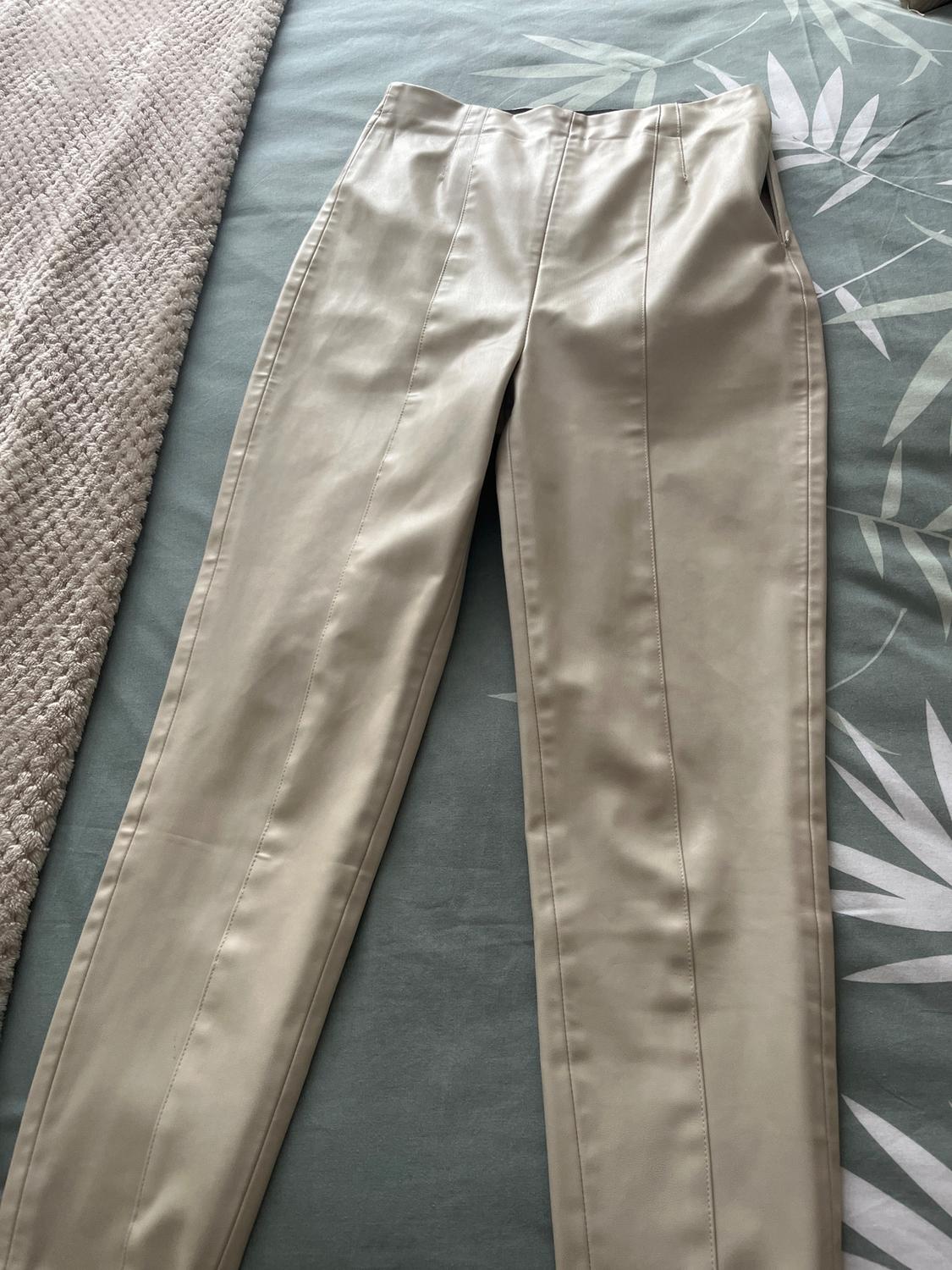 High-waisted beige trousers with visible seams, placed on a bed with a grey bedspread and a leaf-patterned sheet.