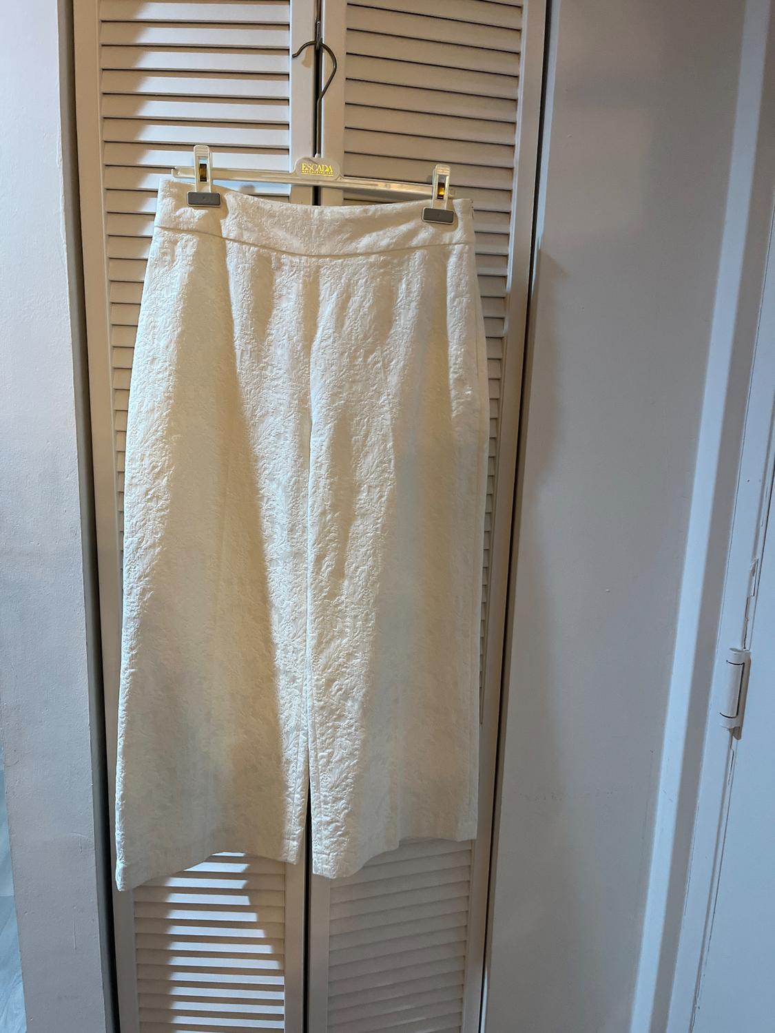 Textured white trousers hanging on a gold hanger with the "ESCADA" brand, in front of shuttered doors in an indoor setting.