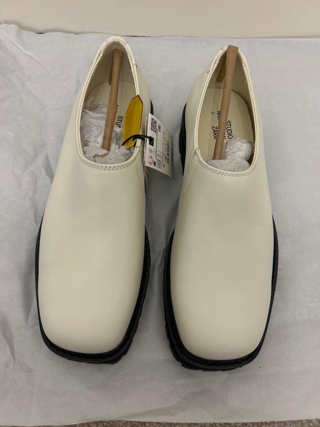 White square-toe shoes with black soles, stuffed with paper and wooden supports, on white paper. Label visible.