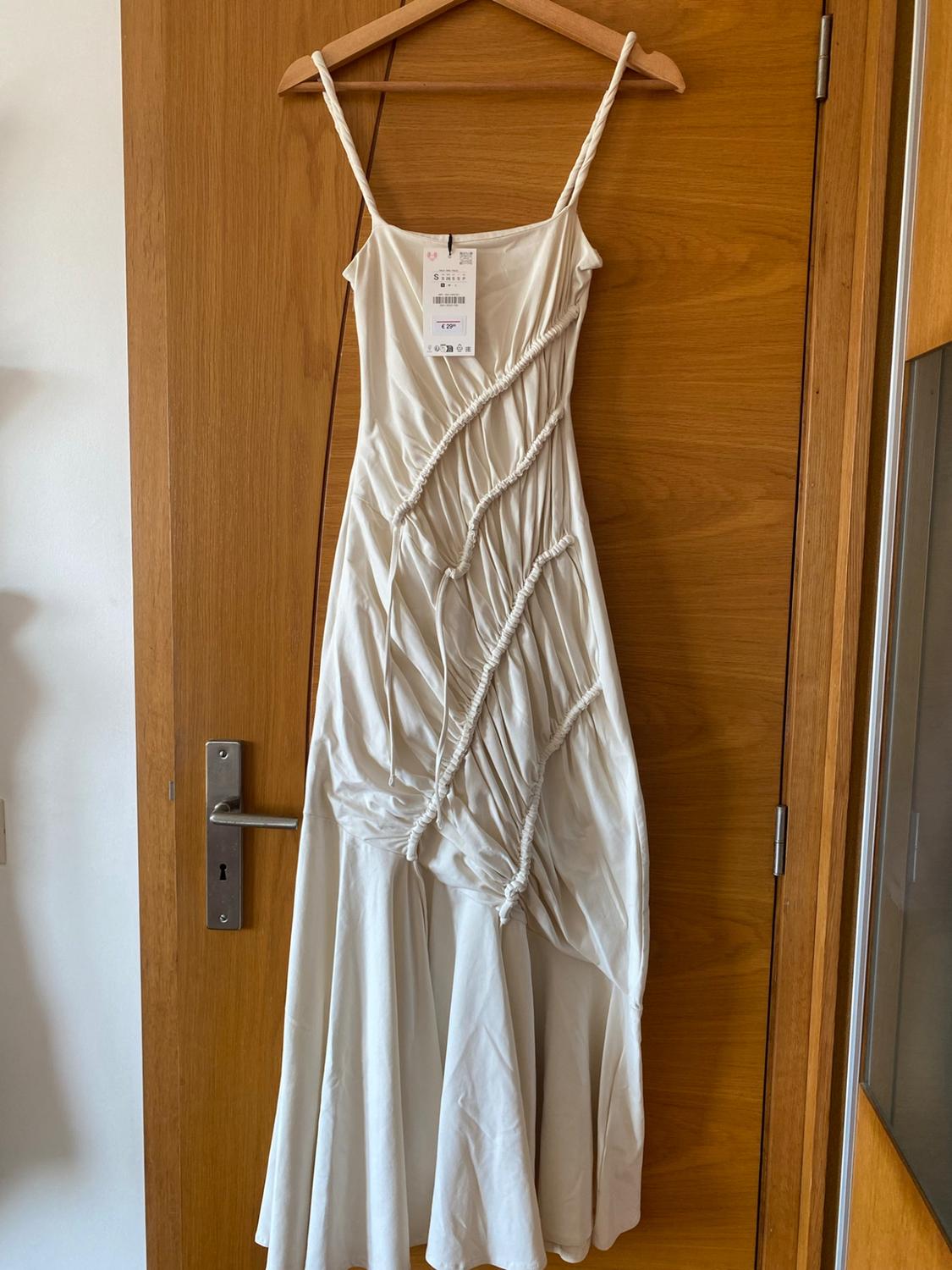 Cream long dress with braided straps and diagonal gathered design, hanging on a hanger over a wooden door.