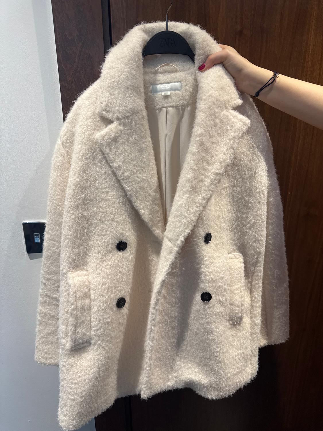 A white wool coat with black buttons is hanging on a black hanger, held by a person's hand. The background is wooden.
