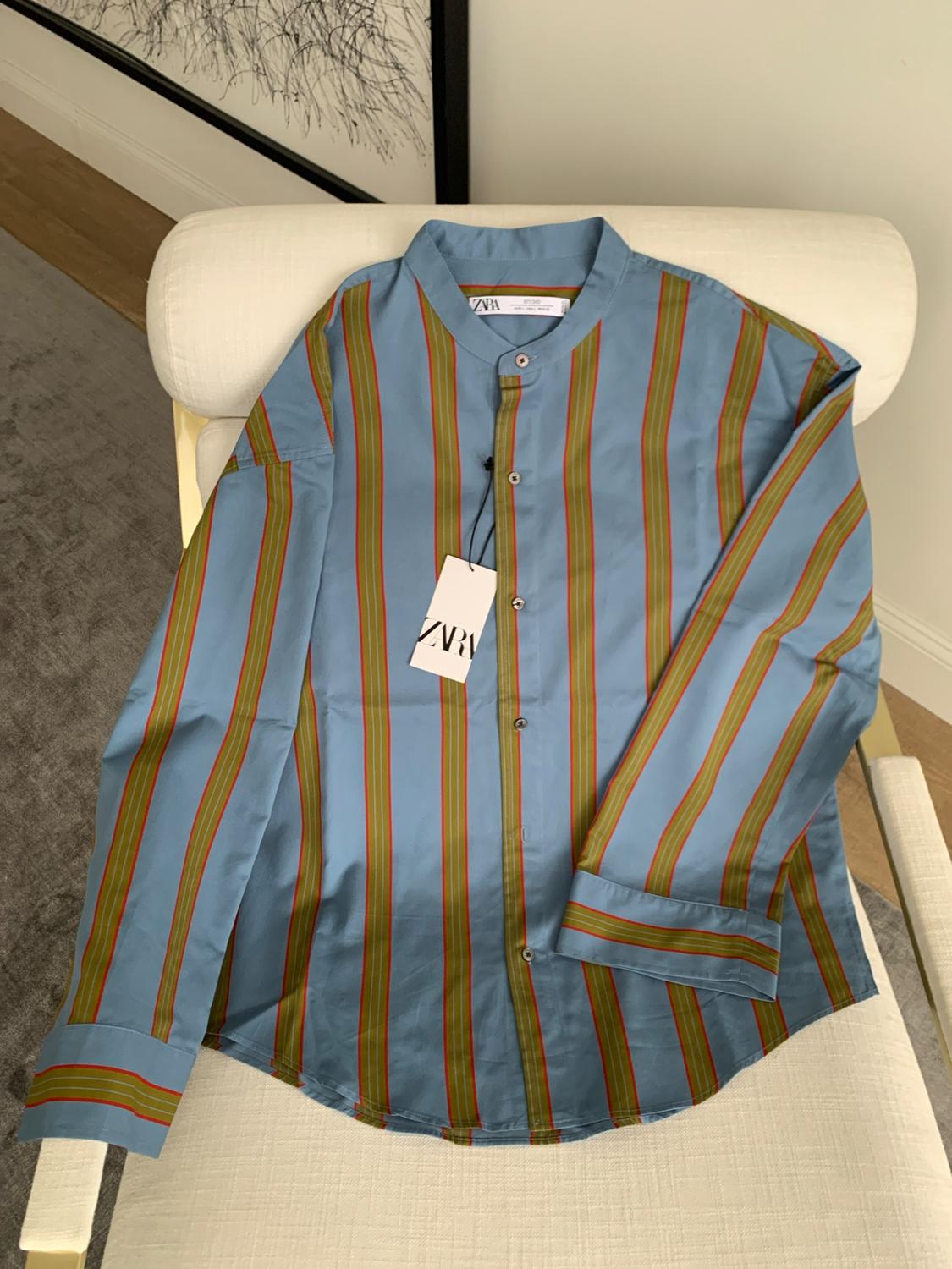 Blue shirt with vertical stripes and a mandarin collar on a white chair. Label with the text ZARA. Relaxed indoor style.