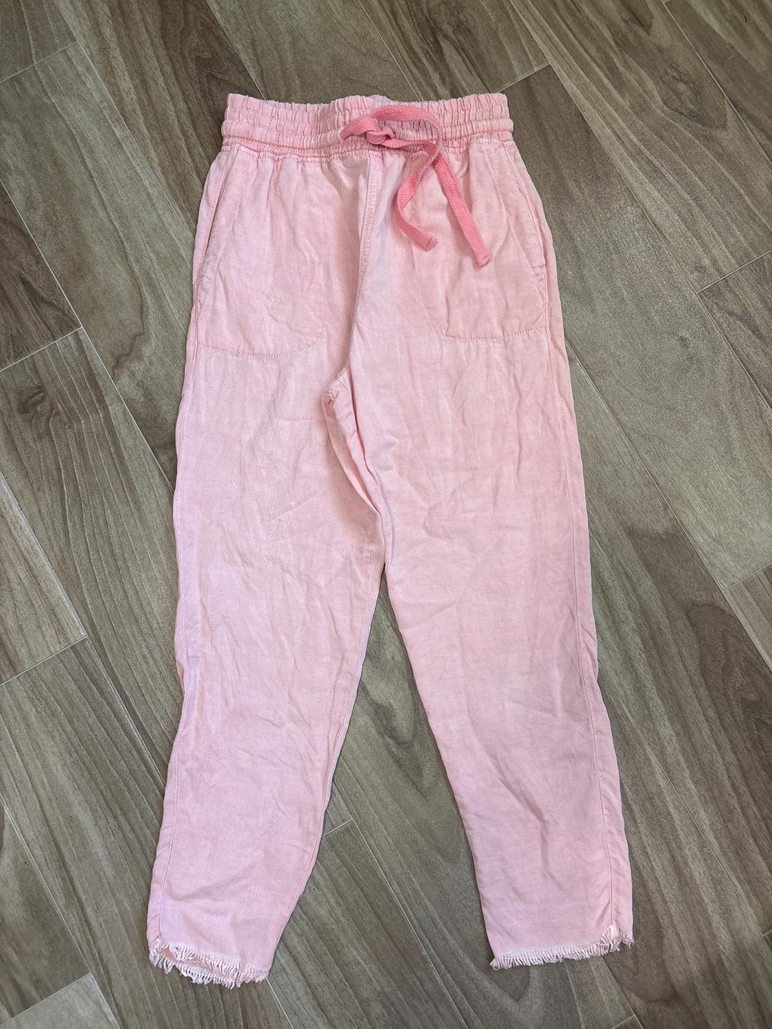 Light pink fabric pants with elastic waistband and drawstring, front pockets, and frayed hem, on a wooden floor.