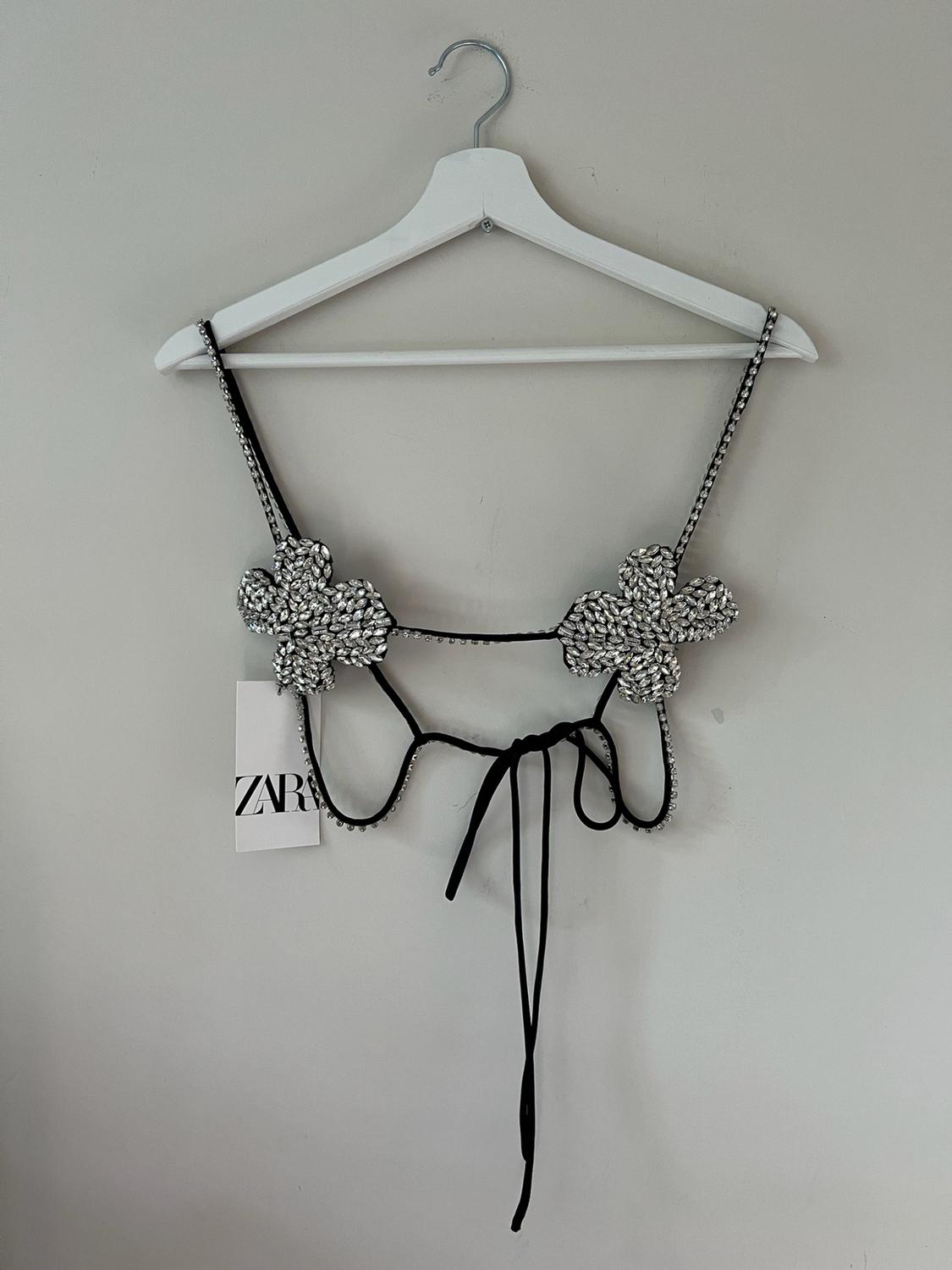 Black camisole top with silver rhinestone flowers on a white hanger against a light background. Label with the text ZARA.