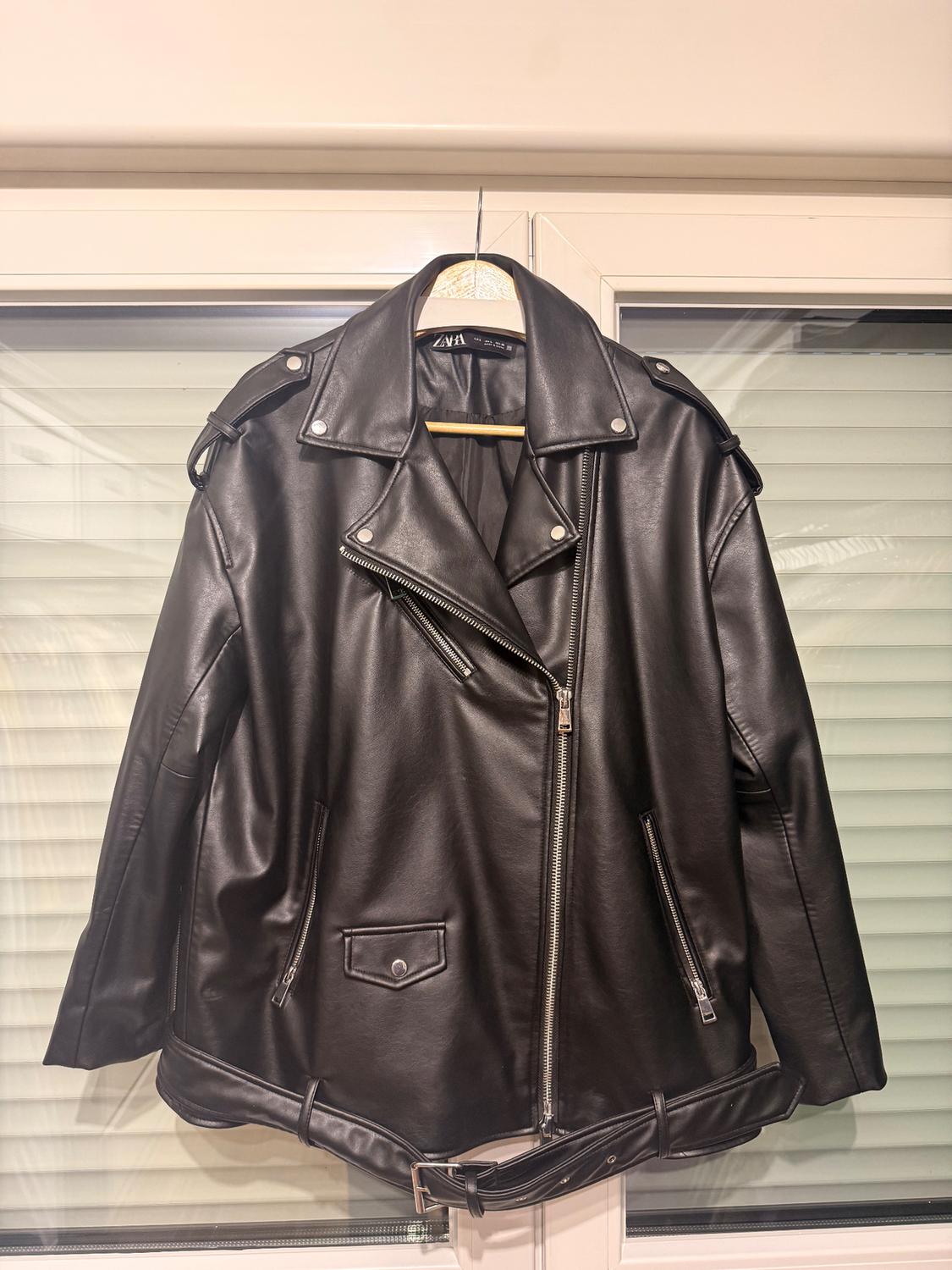Black leather jacket hanging on a hanger in front of a window. It has zips and a belt at the bottom.