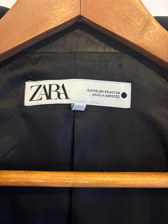 Label from a black garment hanging on a wooden hanger. Visible text: "ZARA EUR XS USA XS MEX 24 MADE IN MOROCCO".
