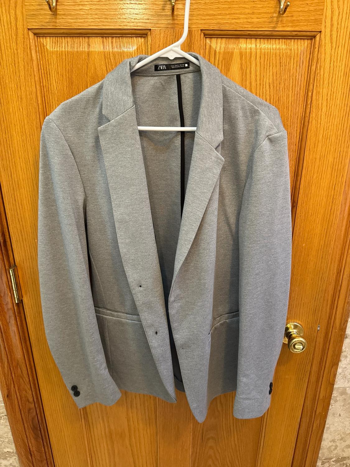 Grey blazer with a ZARA label, hanging on a white hanger over a wooden door with a gold handle in a relaxed setting.