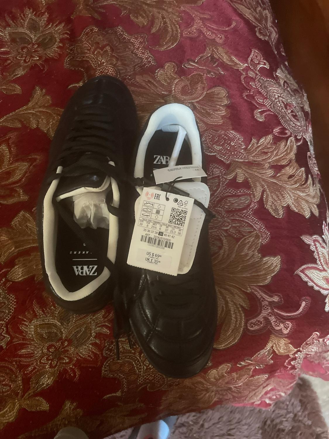 Black ZARA sneakers with white interior on printed red fabric. Label with visible text: ZARA, US $ 69.90, UK £ 35.99.