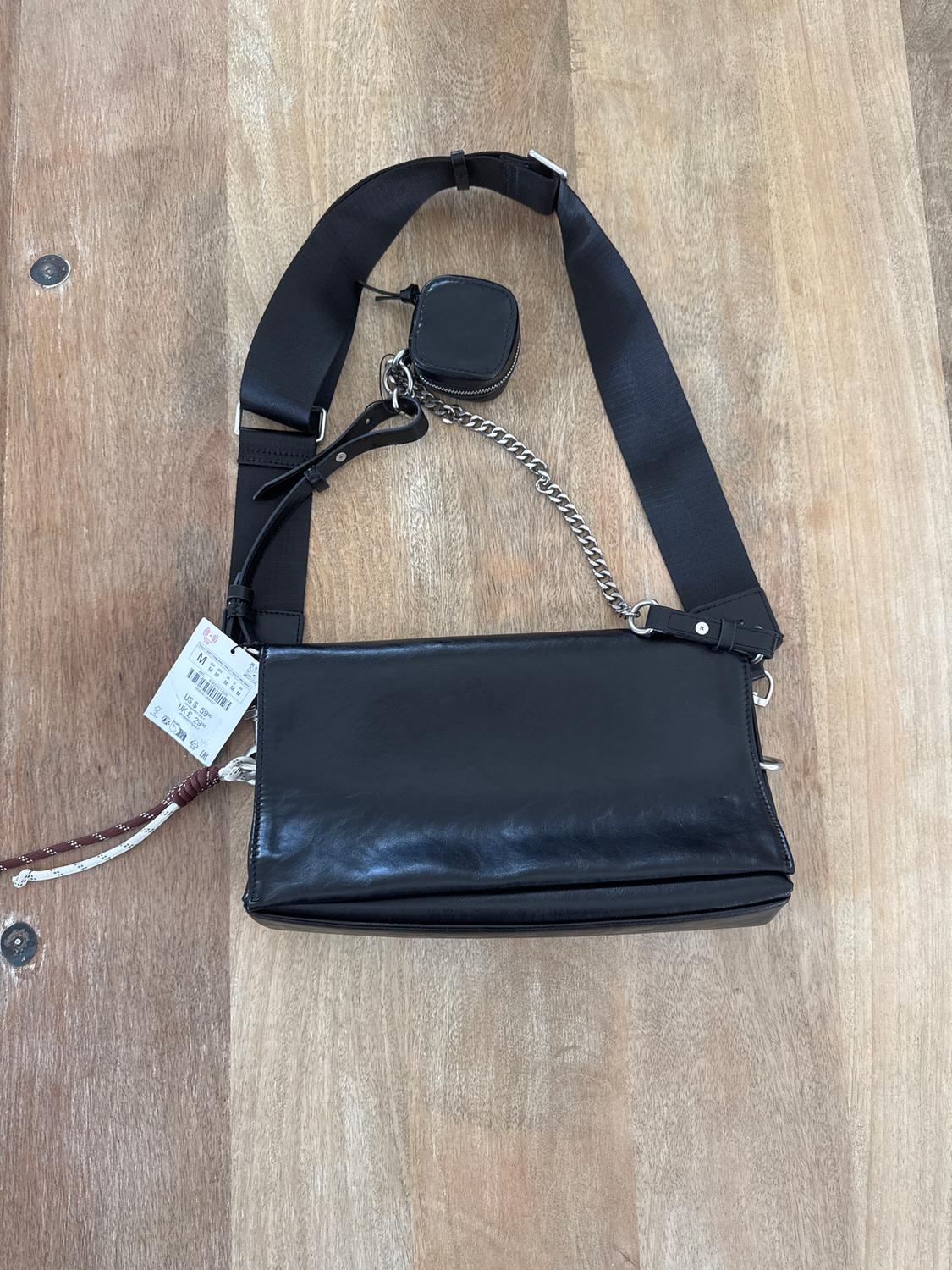 Black rectangular handbag with a wide strap and metal chain on a wooden table. White tag hanging from the left side.
