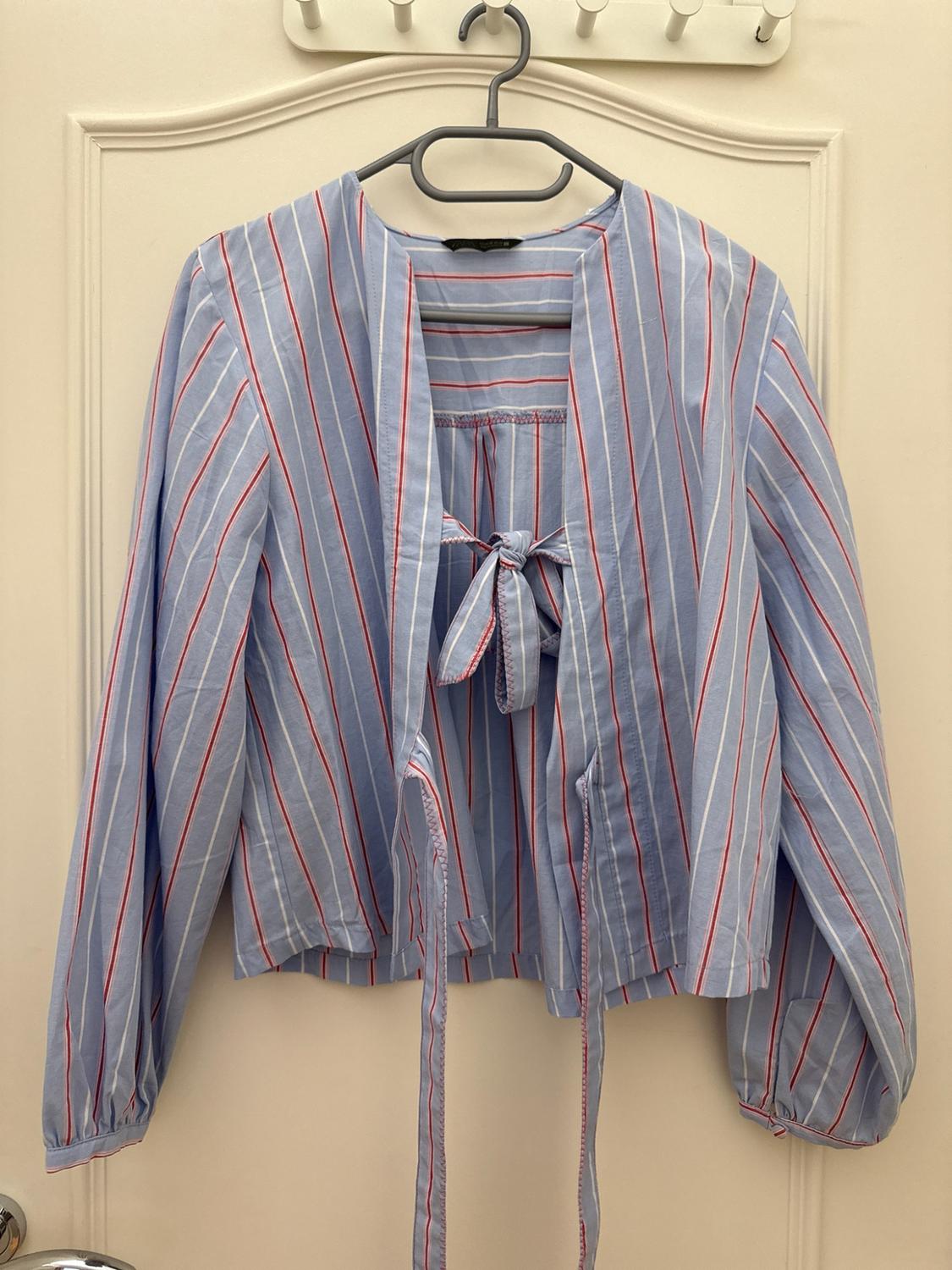 Light blue blouse with red and white stripes, long sleeves and bow detail. It is hanging on a hanger over a white door.