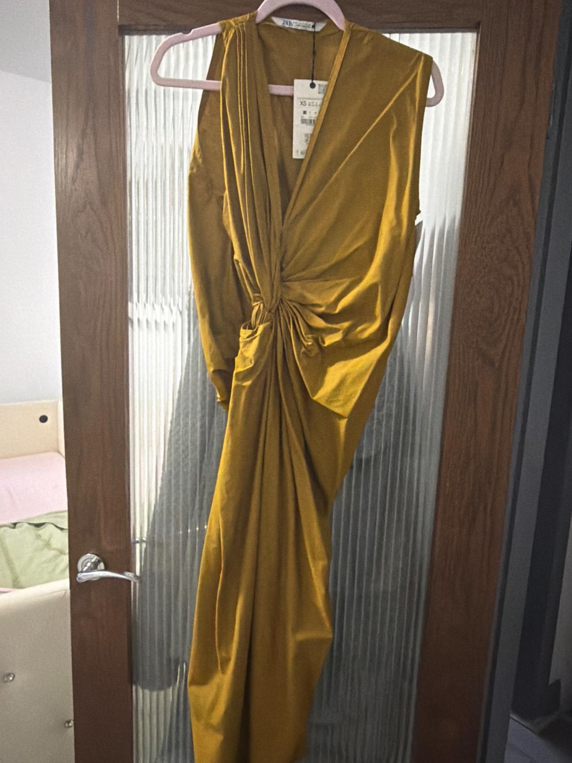 Draped gold dress hanging on a pink hanger over a textured glass door. Label visible on the collar.
