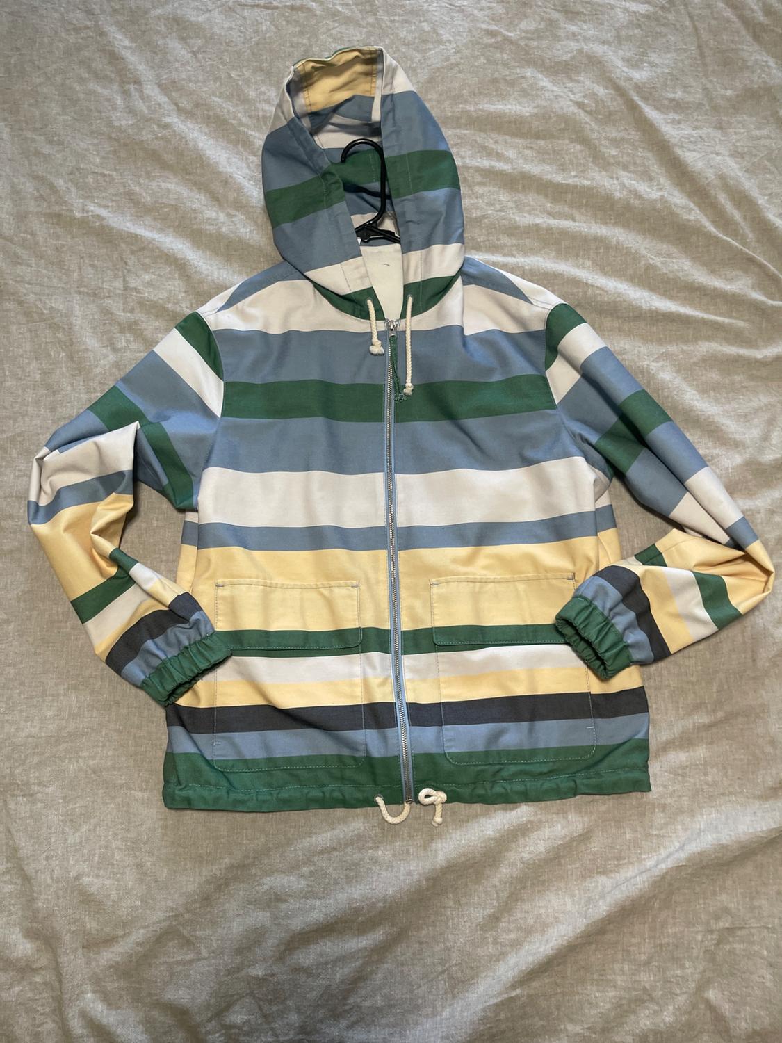 Hooded sweatshirt with horizontal stripes in shades of blue, green, white, and yellow, on a grey background.
