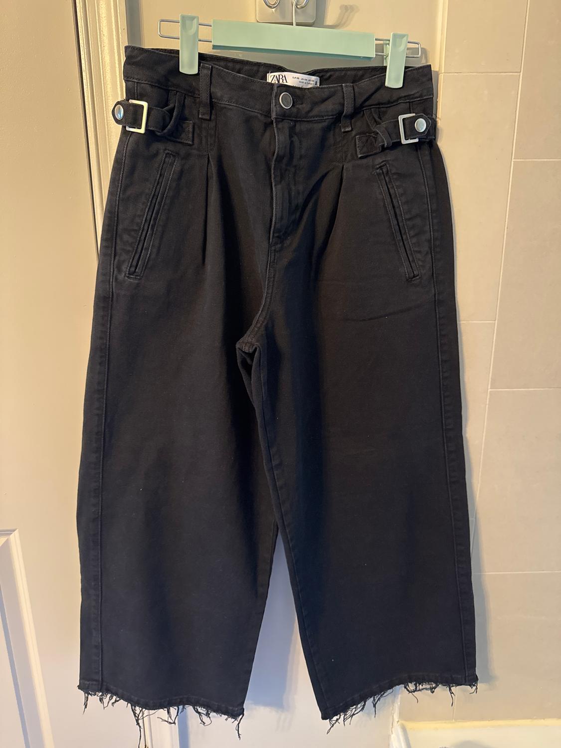 Black trousers hanging on a green hanger, with side adjusters and a frayed hem. Visible label at the waist.