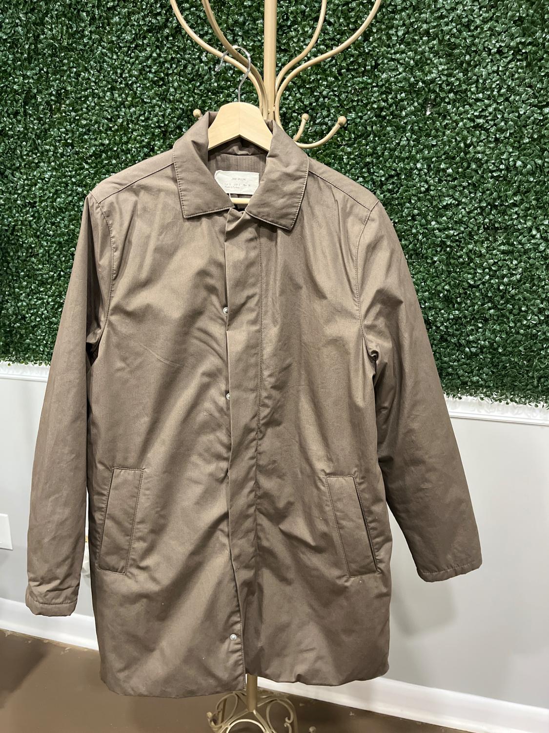 Brown coat hanging on a golden rack in front of a green leafy background. The coat has pockets and front buttons.