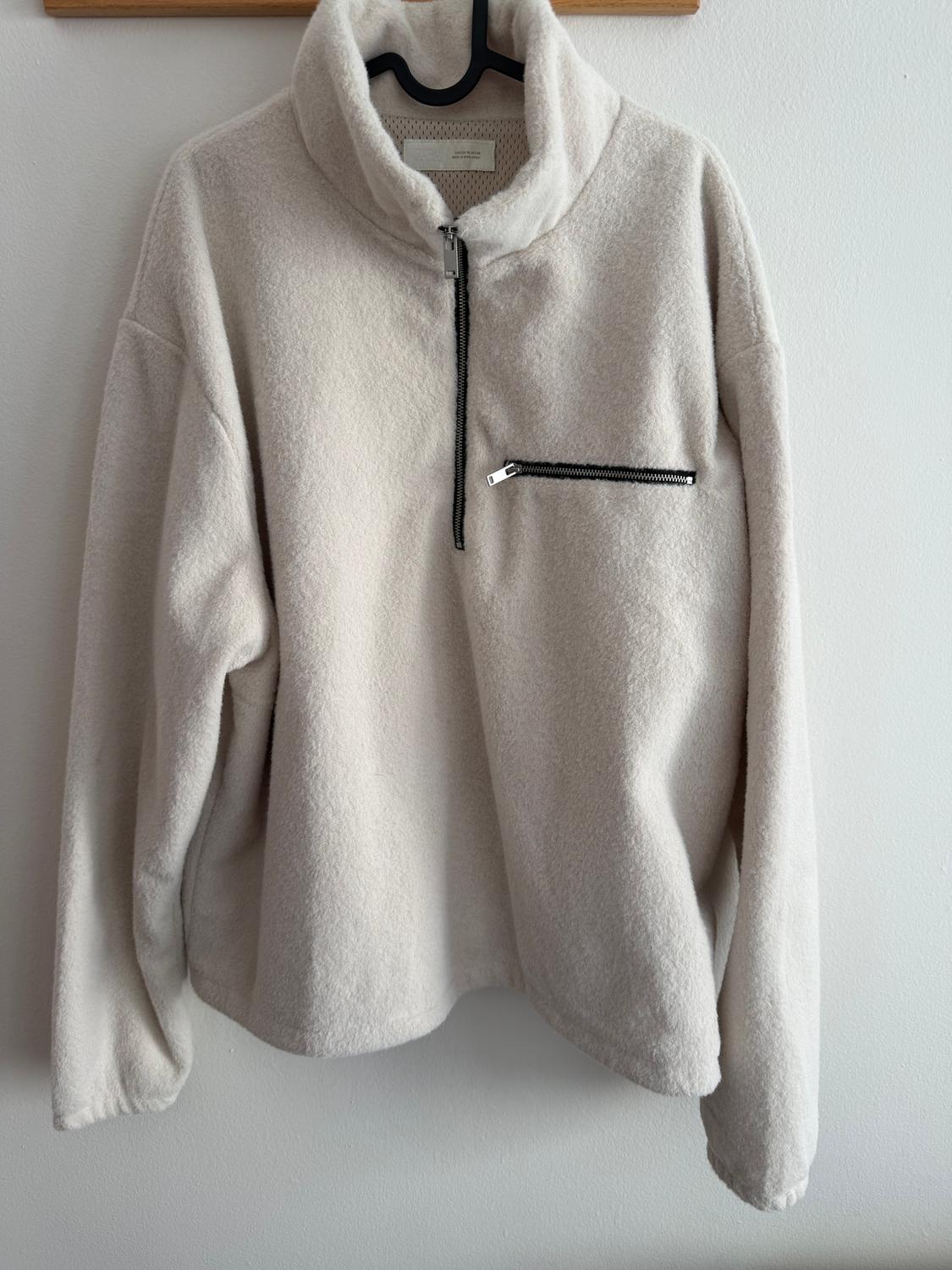 Beige wool jumper with a zip on the chest, hanging on a black hanger against a white wall.