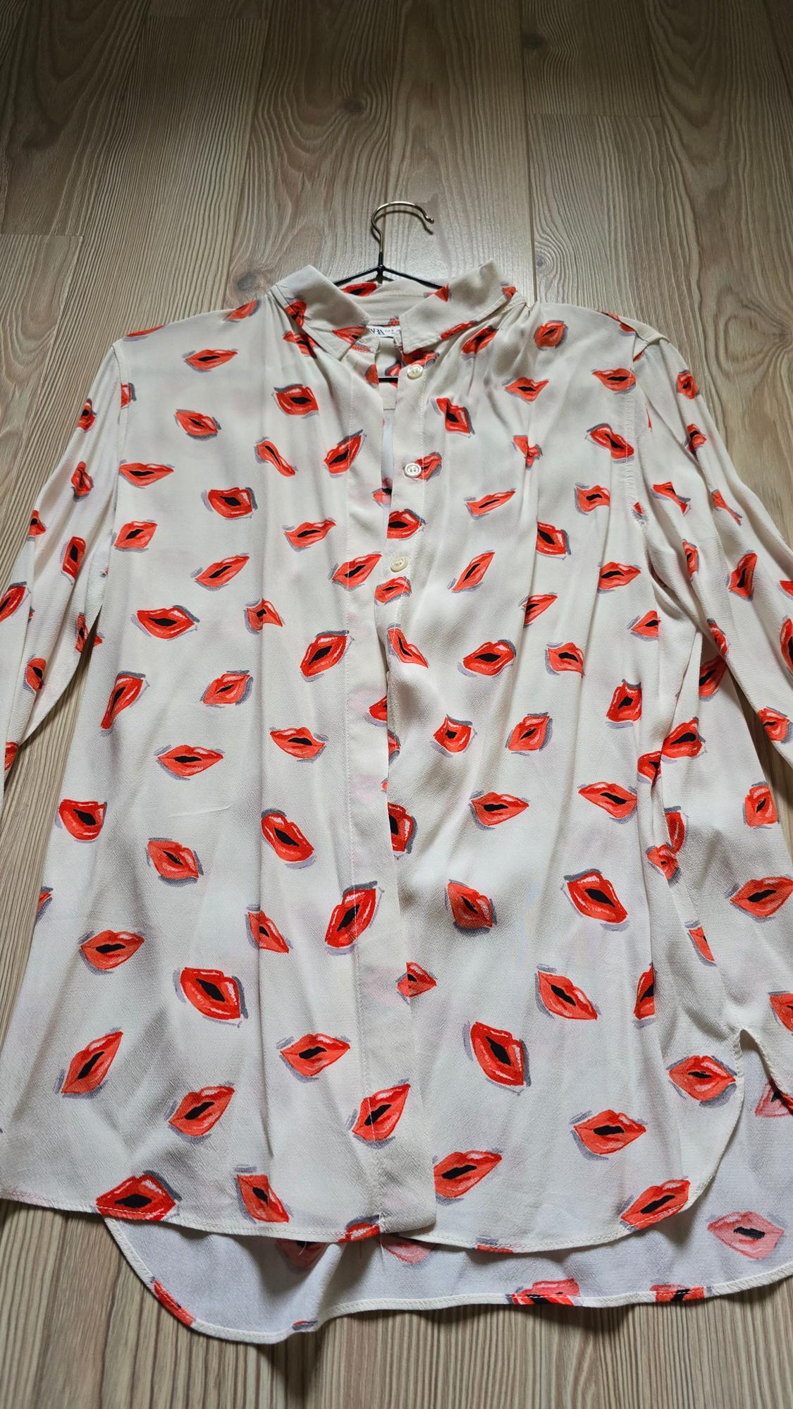 A white blouse with a red and black lip design, hanging on a hanger over a light wooden floor.