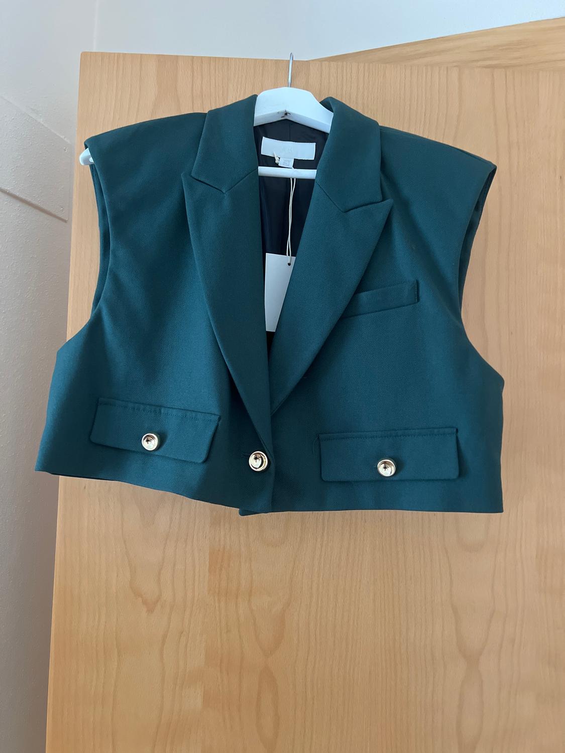 Dark green sleeveless waistcoat hanging on a white hanger over a light wooden door, with gold buttons.
