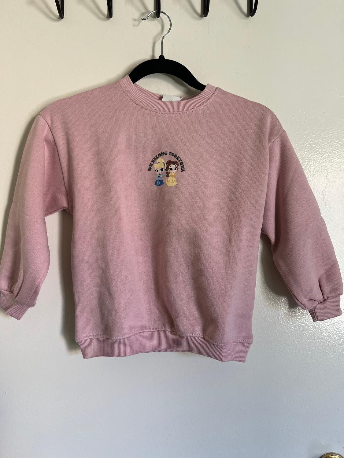 Pink sweatshirt hanging on a black hanger, with illustrations of two characters and the text "WE BELONG TOGETHER" on the chest.