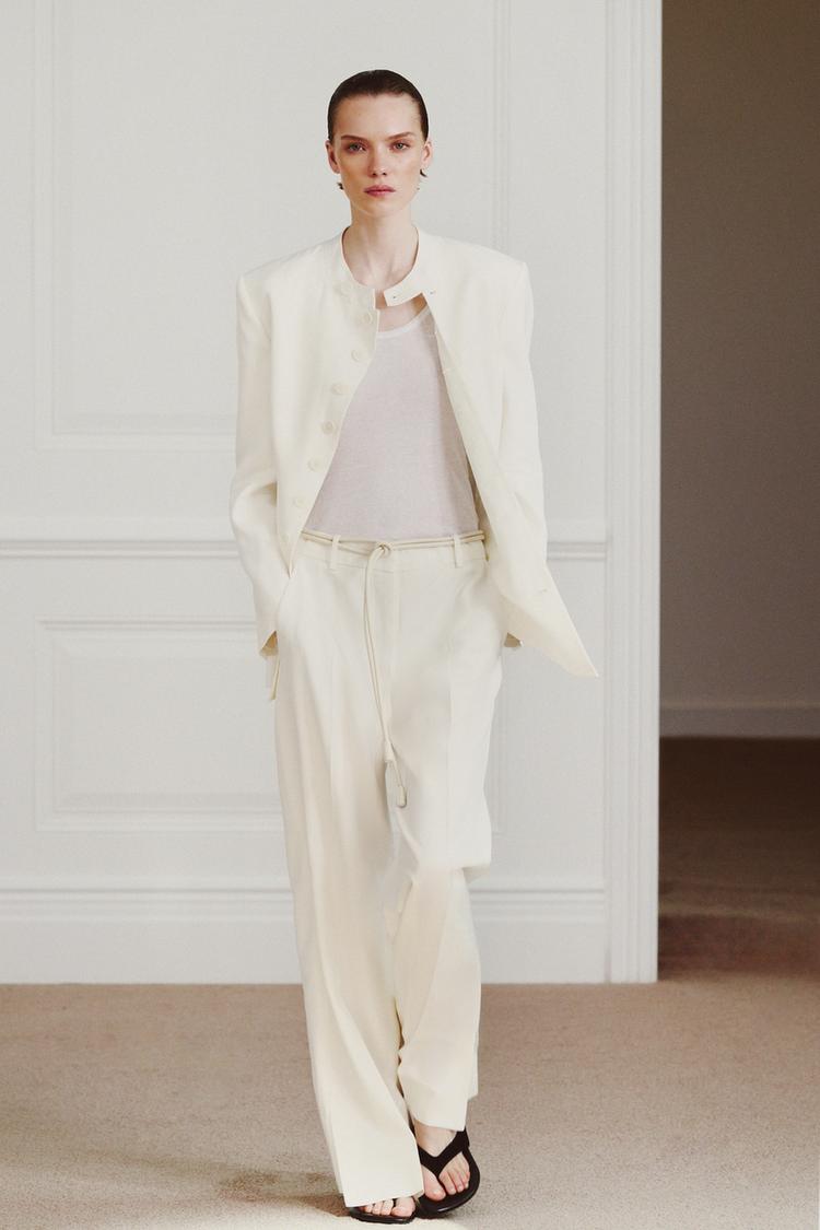 Komplet rovné krémové bundy a kalhot s bočními kapsami a asymetrickým lemem. Pod ním bílé tričko.