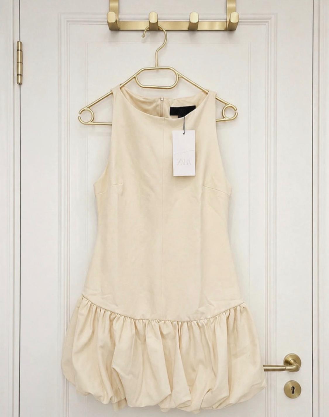 Short beige sleeveless dress with a bubble hem, hanging from a gold hanger on a white door. Label featuring the text ZARA.
