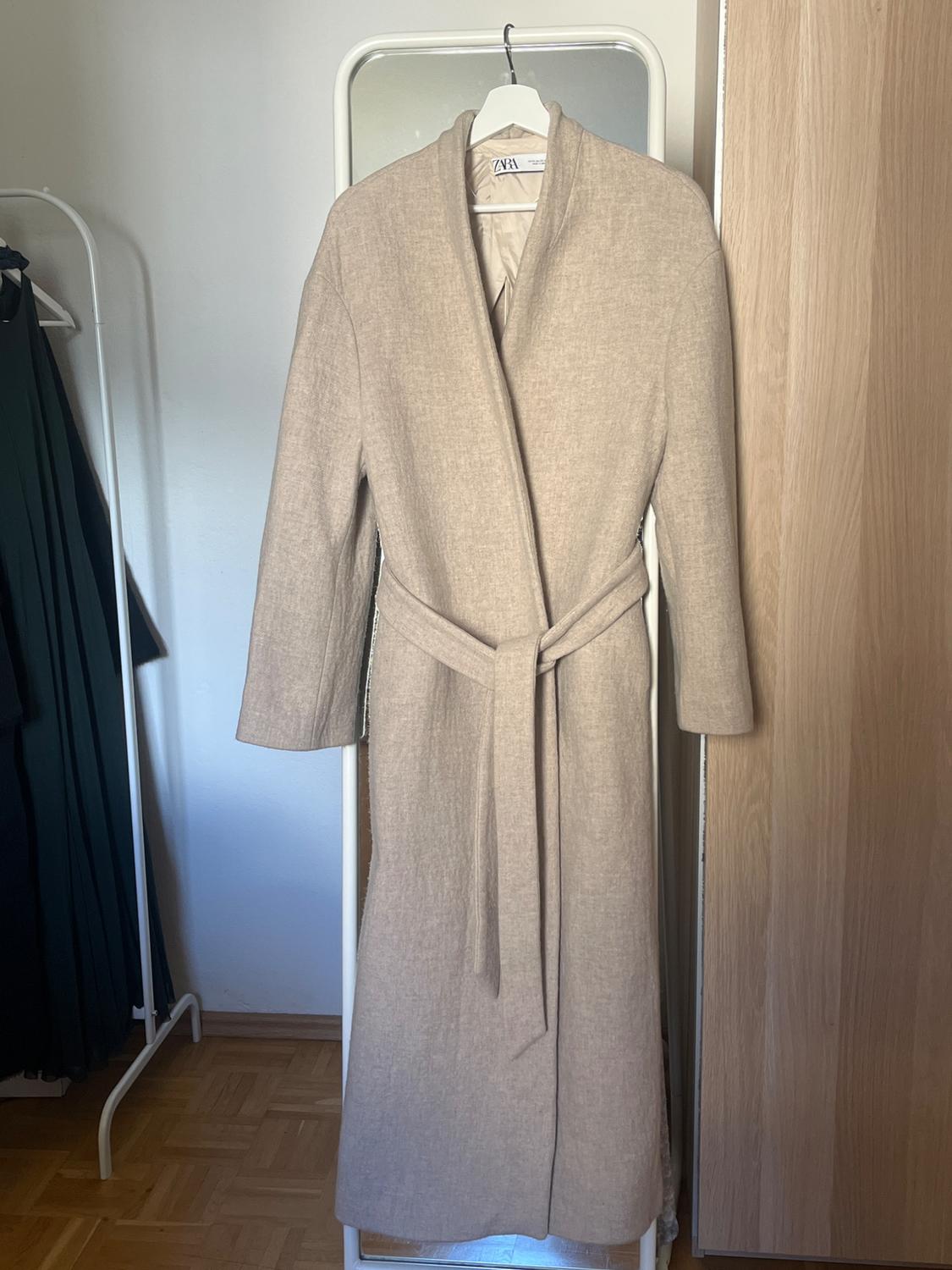 Beige coat hanging on a white hanger in front of a mirror. Background with white wall and light wooden door. Visible label: Zara.