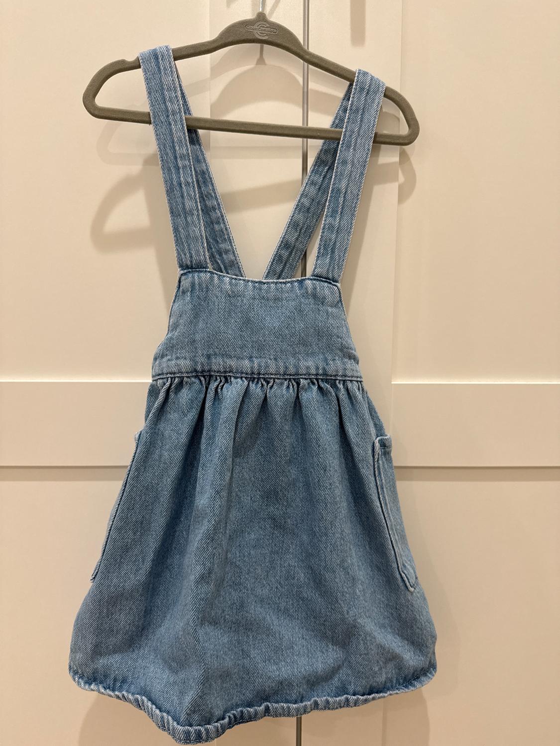 Light blue denim dress with wide straps, hanging on a grey hanger against a backdrop of white doors.