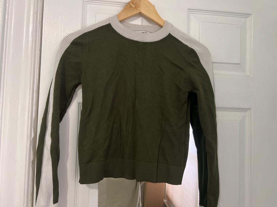 Olive green sweater with long sleeves and white details, hanging on a wooden hanger in front of a white door.
