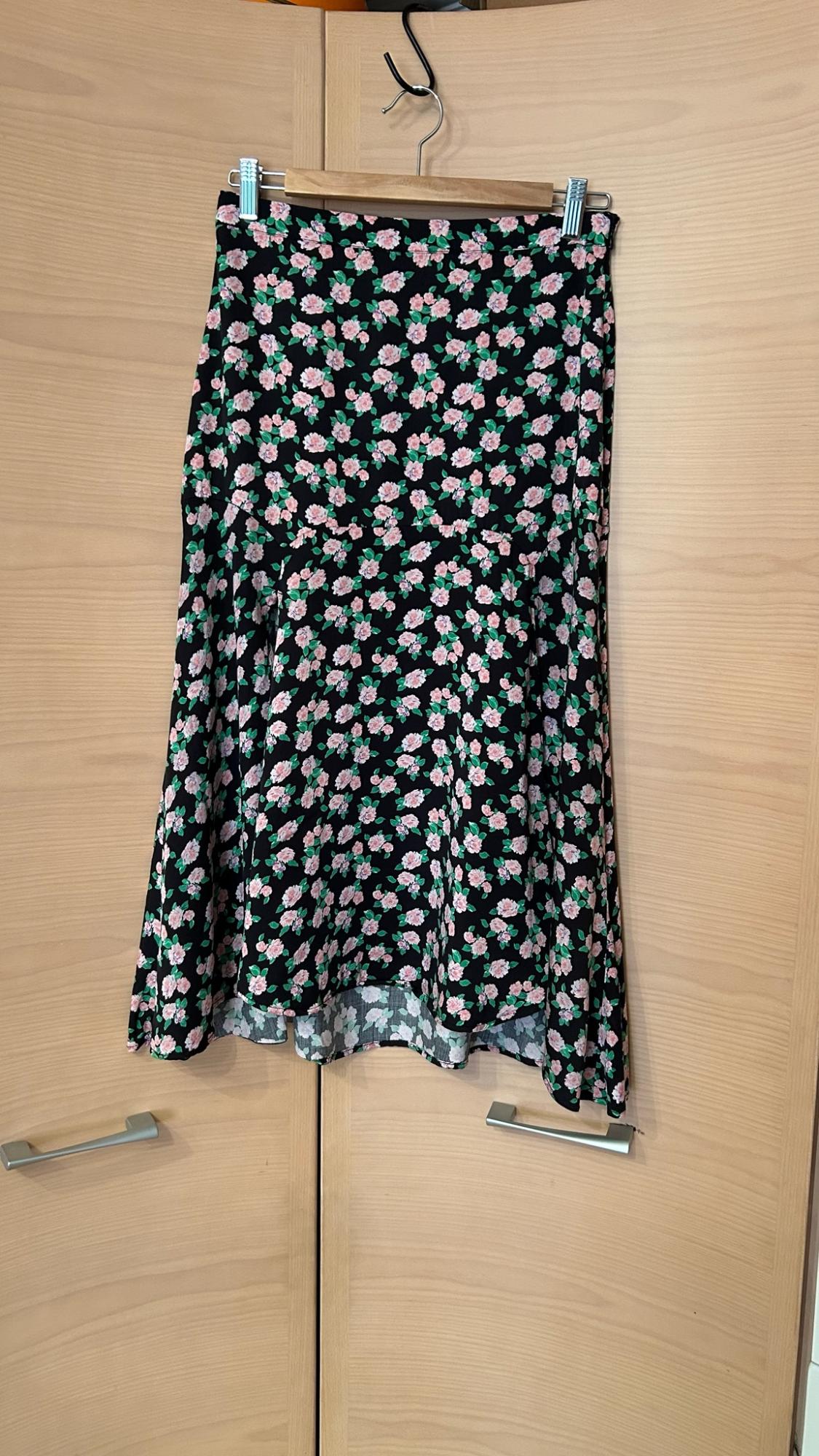 Black skirt with a pink and green floral print hanging on a hanger in front of a light wood wardrobe.