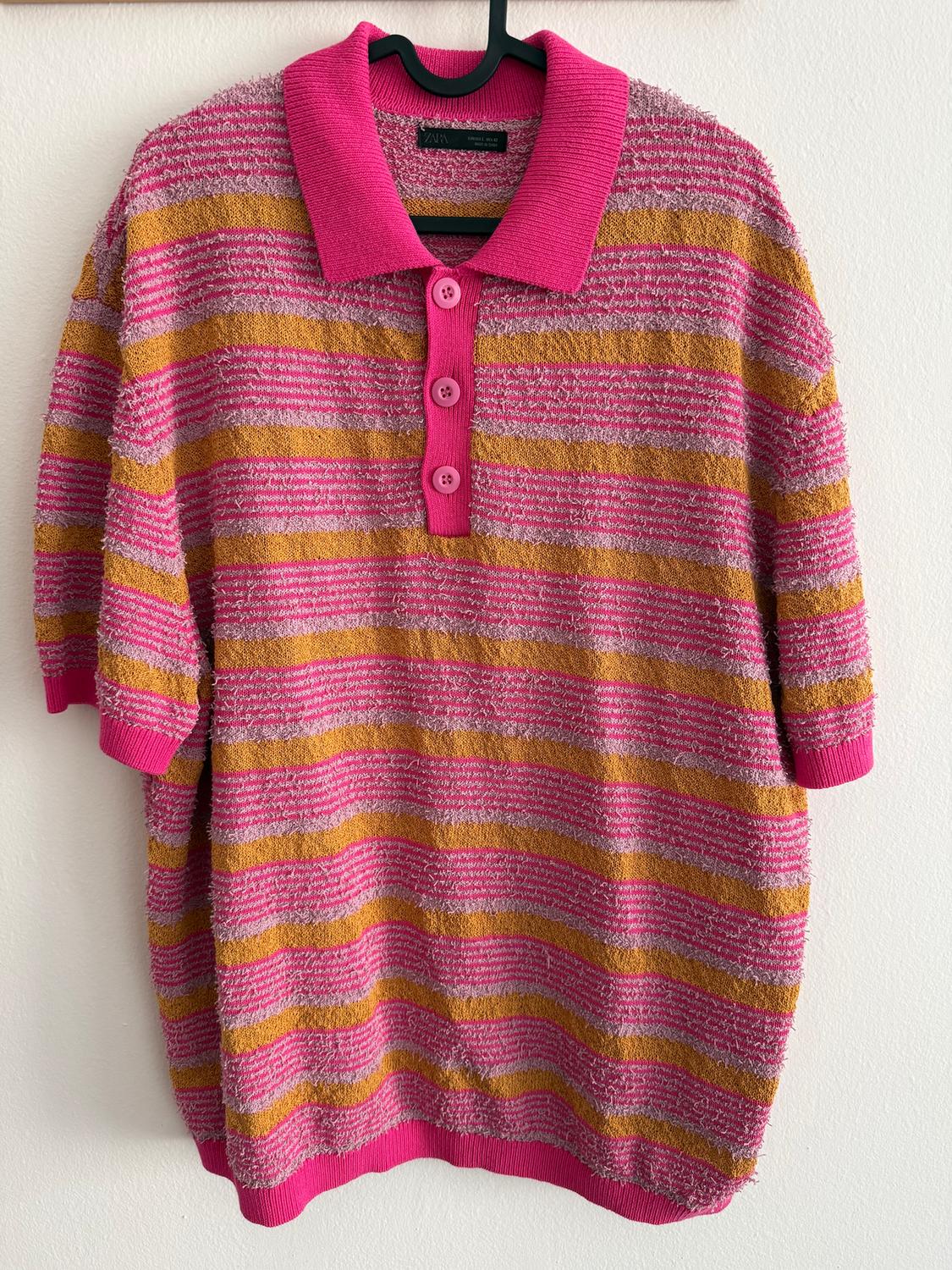 Pink and mustard striped polo neck jumper with pink buttons, hanging on a black hanger against a white wall.