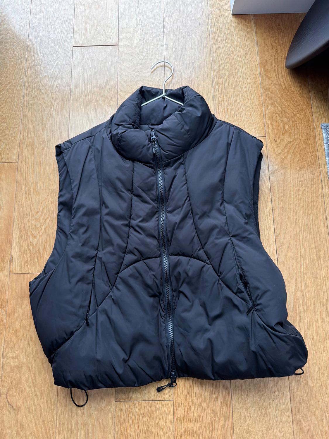 Black padded vest with zipper, hanging on a hanger over a light wooden floor.