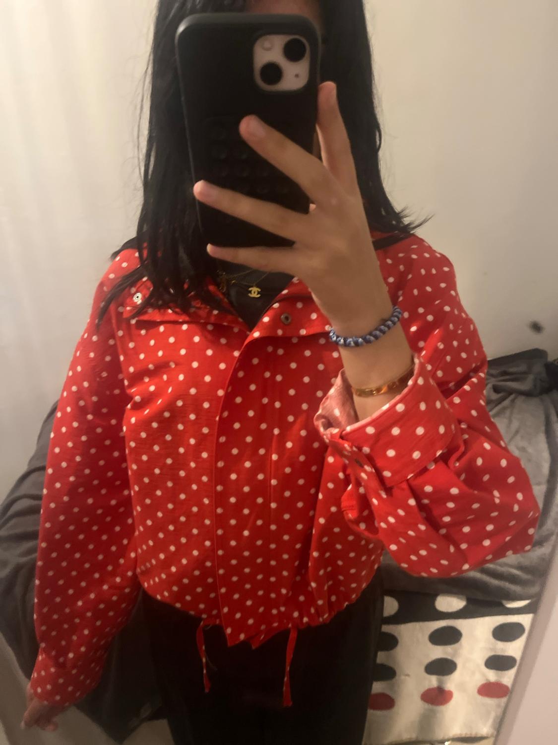 Mirror selfie. Person with a black mobile phone wearing a red jacket with white spots and dark trousers in a casual setting.