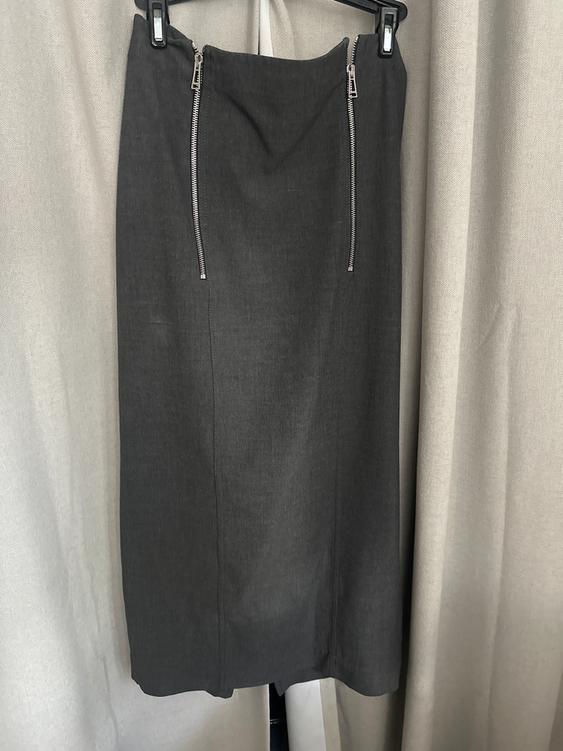 Gray skirt with two vertical zippers at the top, hanging on a hanger against a background of beige curtains.