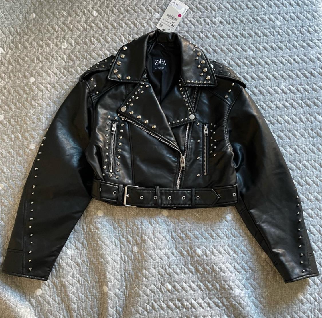 Black faux leather jacket with silver studs and belt, on a gray quilt. Visible label with text "ZARA".