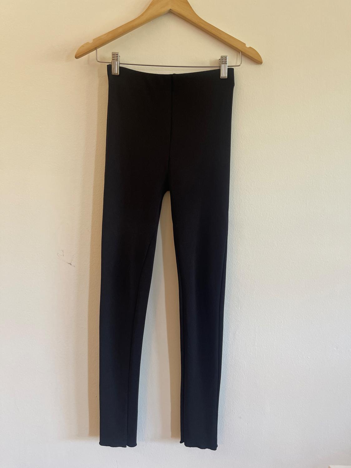 Slim-fit black trousers hanging on a wooden hanger with clips against a plain white wall background. Minimalist style.