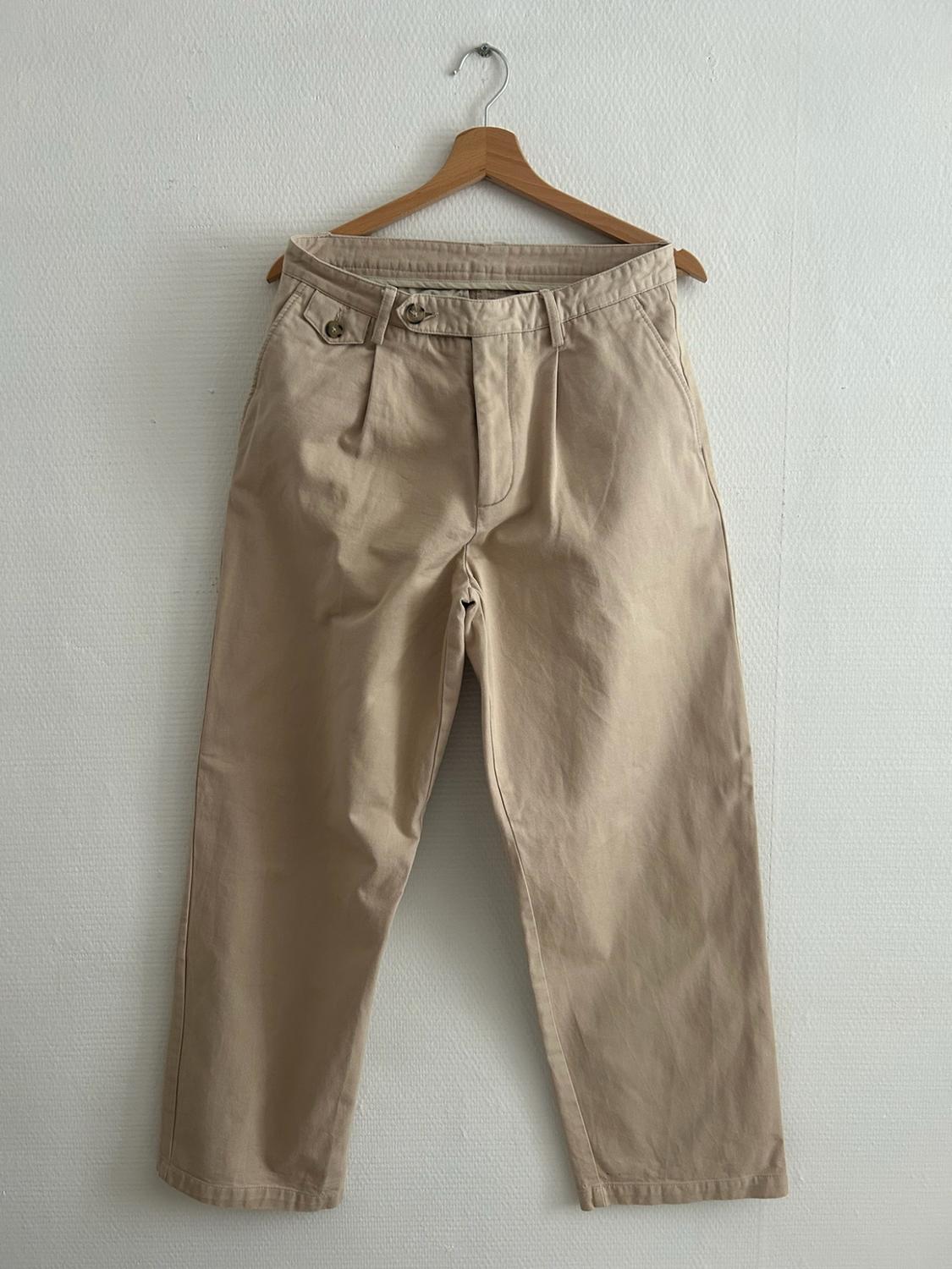 Beige trousers with pleats and a button fastening, hanging on a wooden hanger against a white wall.