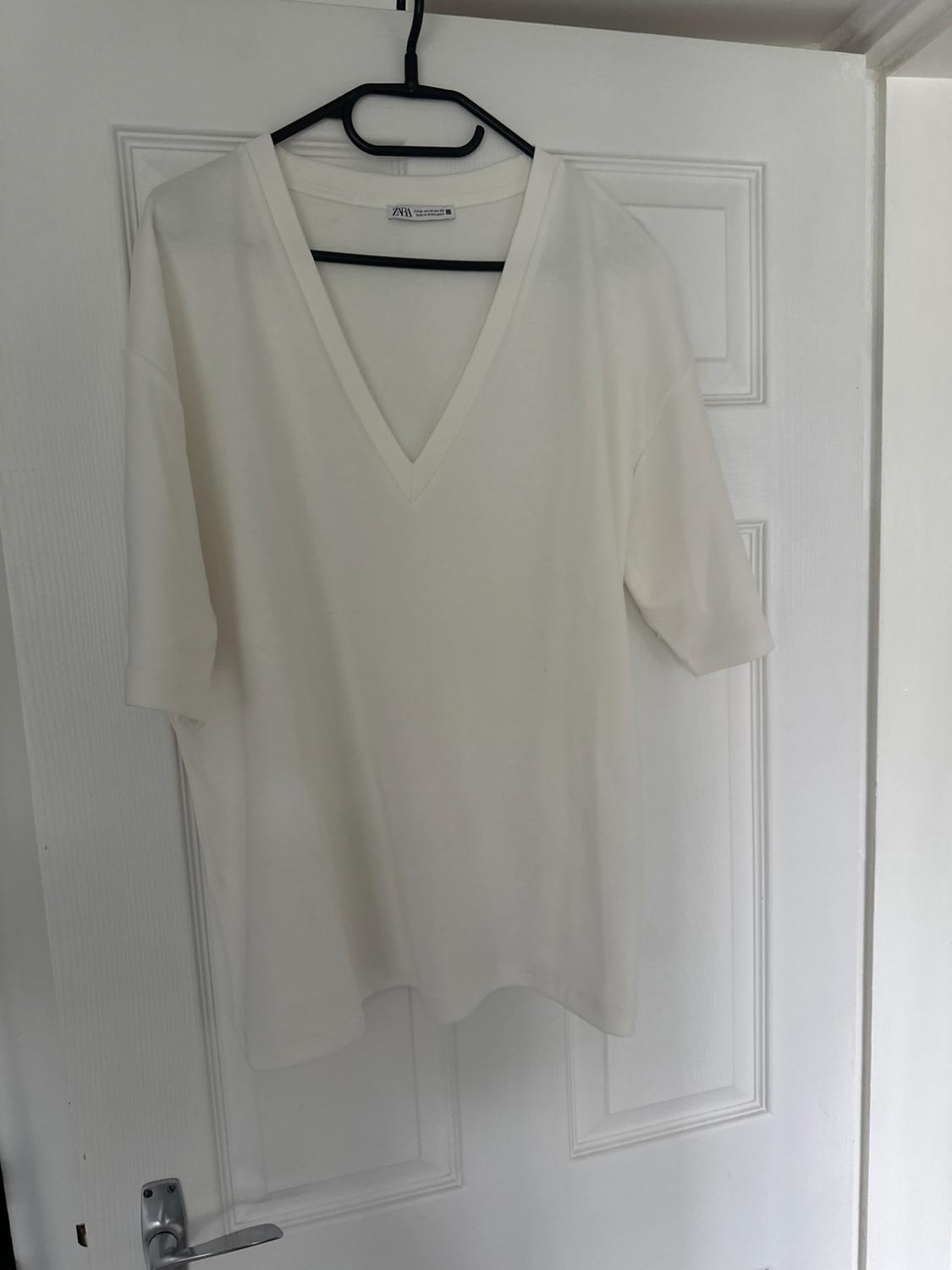 White short-sleeved V-neck T-shirt hanging on a black coat hanger over a white door. Label visible: "ZARA".