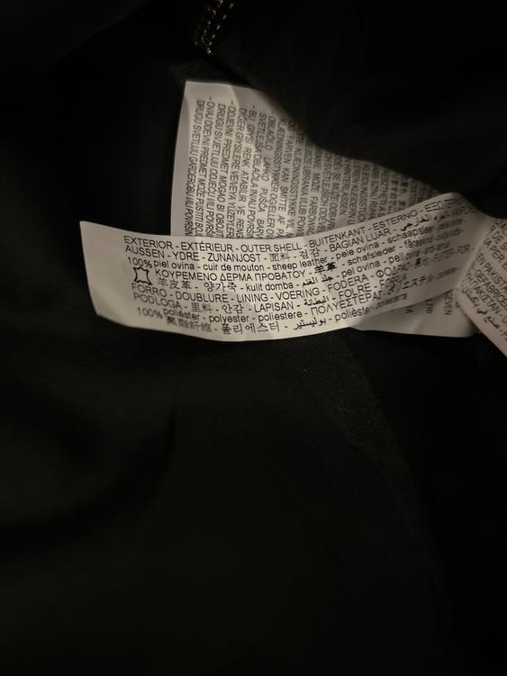 Multi-language clothing label on black fabric, indicating "100% sheepskin" and "100% polyester" for outer and lining.