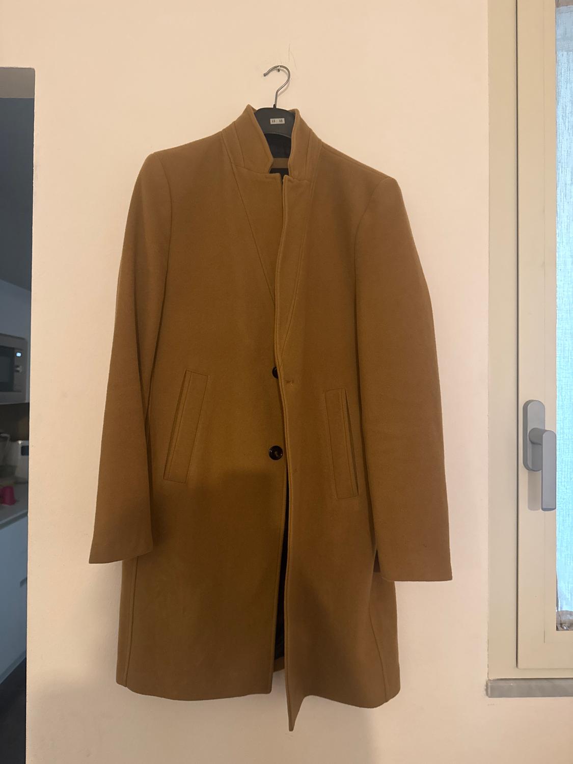 Brown coat hanging on a hanger against a white wall, with two front pockets and a visible button.