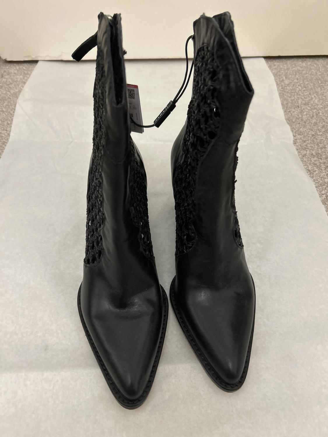 Black pointed boots with lace details on the sides, on white paper on a grey floor. Visible label on one boot.