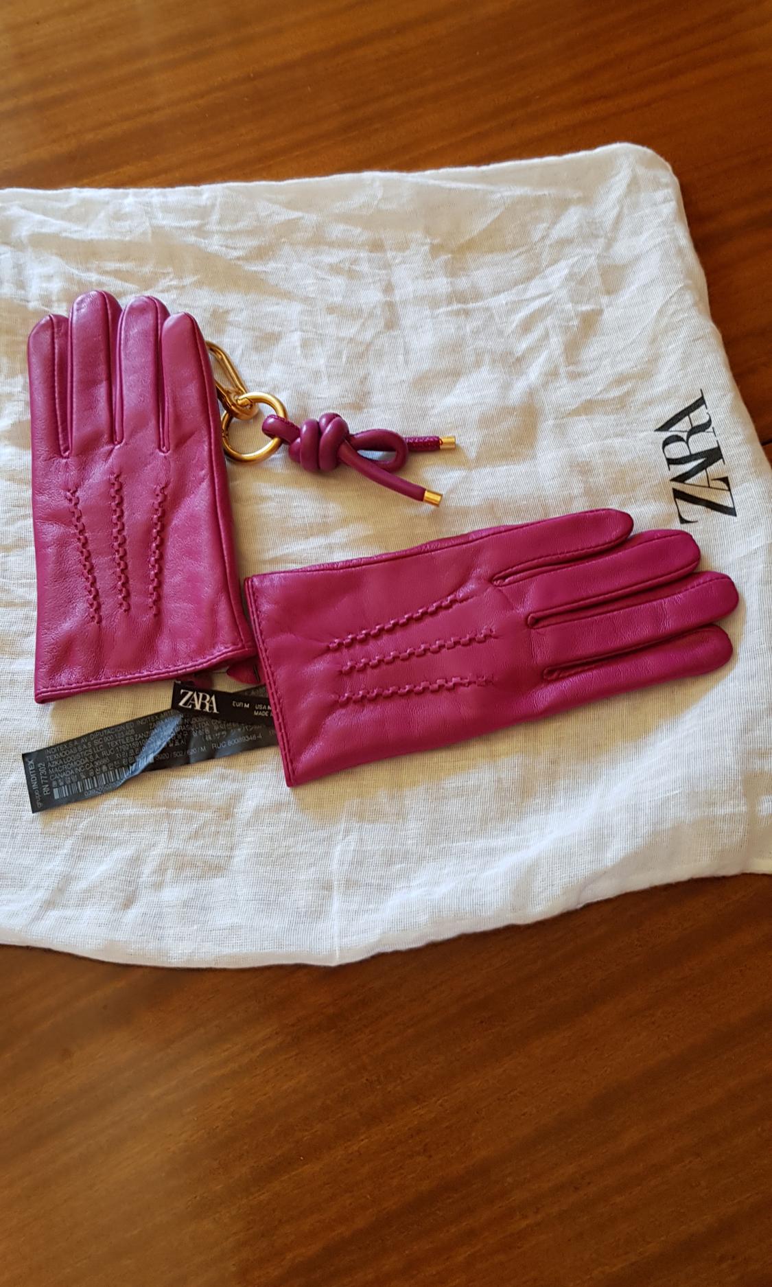 Fuchsia gloves on white fabric with a label and gold keyring. Visible text: "ZARA". Brown wooden background.