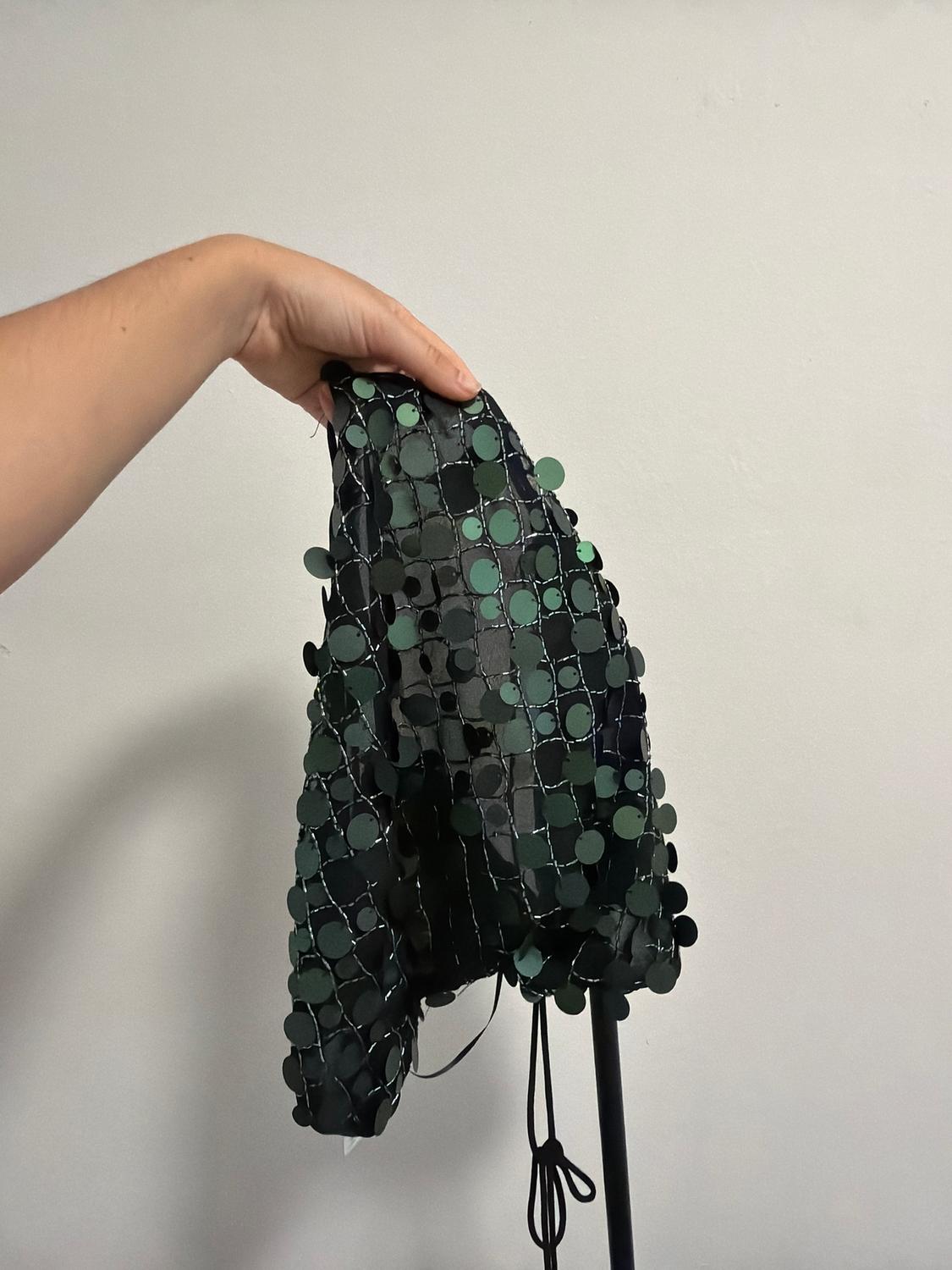 Hand holding black fabric with green sequins on a white background. The fabric has a grid design with shiny circles.