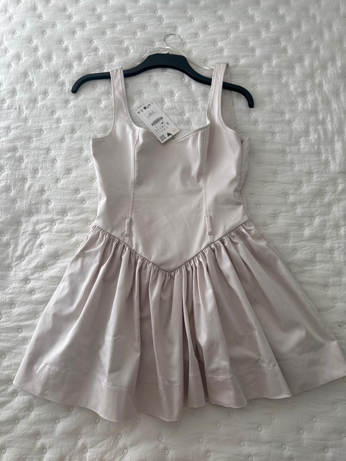 Light pink dress with wide straps and a flared skirt, hanging on a black hanger over a white quilted bedspread.
