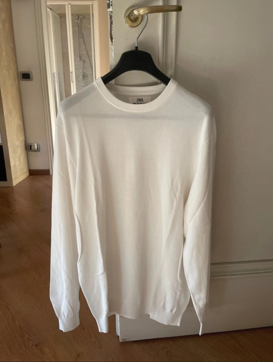 White, long-sleeved jumper hanging on a hanger on a white door handle. Label with ZARA text. Homely setting.